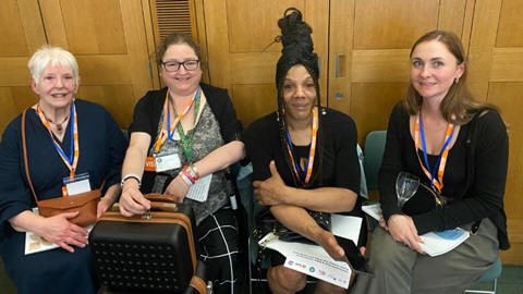 Four women seated at an event, wearing lanyards and holding documents and bags.