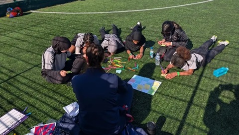 Children taking part in an outdoor activity on an artificial sports pitch, writing ideas on cards while sitting in a group with a facilitator.