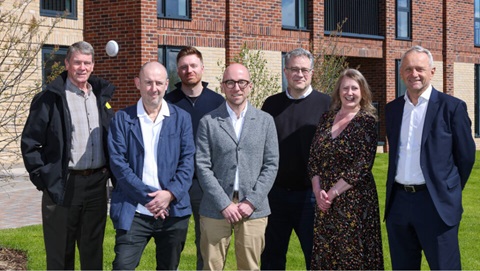 Group of seven adults standing on grass outside a modern brick residential building, posing for a photo.