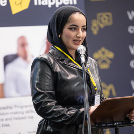 Woman speaks at a podium with a microphone during an event, wearing a black headscarf and leather jacket.