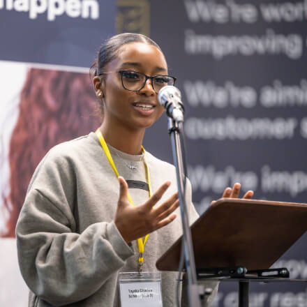 Woman presents confidently at a podium, gesturing while speaking into a microphone