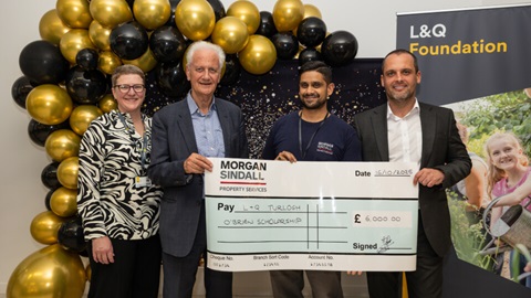 Four people stand smiling with a large presentation cheque for the L&Q Turlough O’Brien Scholarship, in front of gold and black balloon decorations.