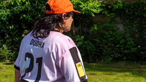 Back view of a Union Manchester FC team member wearing a light purple jersey with "Dudha" and number 21, standing on grass with greenery in the background. On the sleeve, the L&Q logo can be seen.