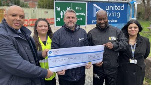 Five people smiling and holding a £1,000 cheque from United Living and L&Q, standing in front of a construction site.