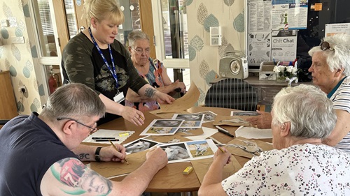 A group of elderly people sitting around a table, working on art projects with a facilitator standing nearby.