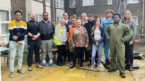 A large group of community members and volunteers standing together in a courtyard with gardening equipment, showcasing teamwork.