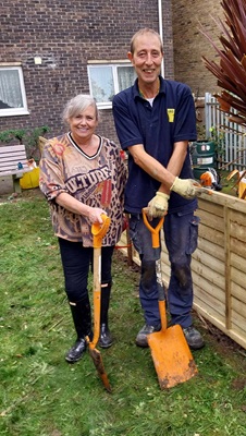 Two smiling people holding orange shovels, standing in a garden area with fencing and tools in the background.