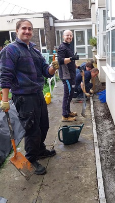 A row of people actively digging and working along a building's pathway, with shovels and a watering can.