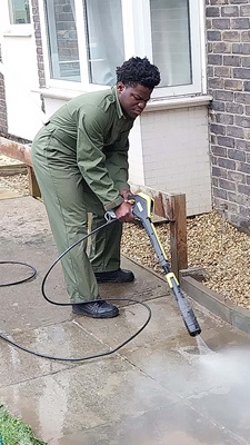 Person in green overalls using a pressure washer to clean a paved area near a residential building.