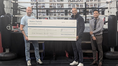 Three people in a boxing gym holding a large check for £5,000, addressed to Carney Community Boxing Club, from Wates Family Enterprise Trust.