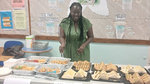 Community volunteer standing behind a table of sandwiches and refreshments at a local event, helping provide food and support for residents.