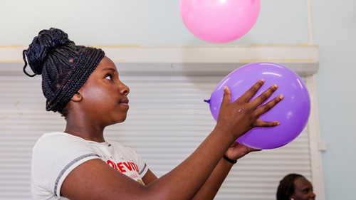 A girl with braided hair and a white T-shirt is holding a purple balloon and looking up at a floating pink balloon indoors.