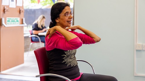 A woman with glasses is seated on a red chair indoors, smiling while doing a stretching exercise with her hands on her shoulders.