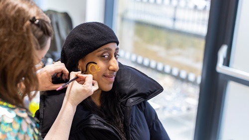 A woman wearing a black coat and black beret is smiling as another person paints a yellow and black design on her cheek.