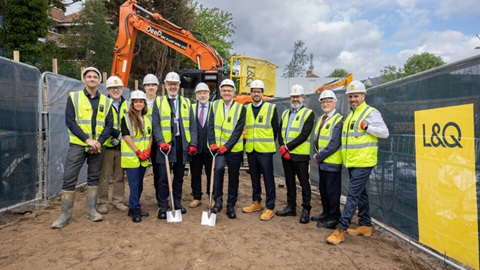 Group of construction and development partners wearing hard hats and high-visibility vests at a ground-breaking ceremony, standing with ceremonial spades on a housing development site.