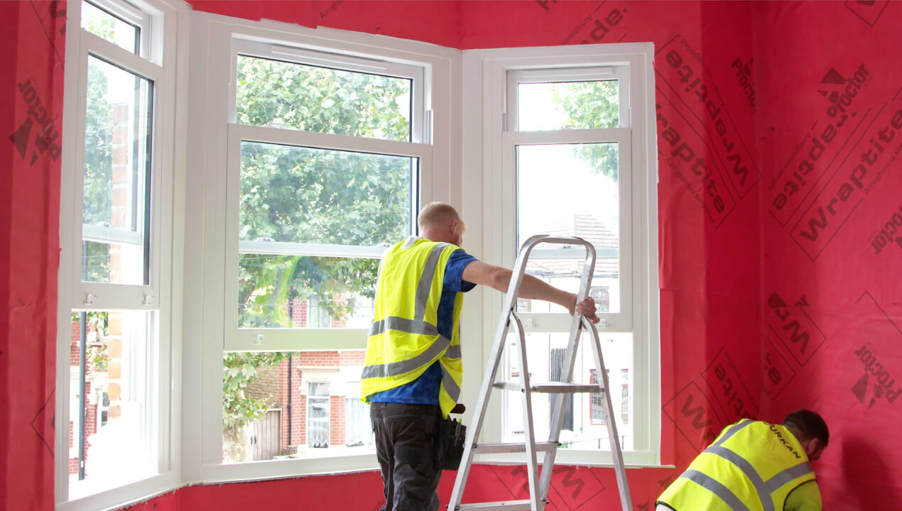 Two workers in high-visibility vests installing insulation in a room lined with red Wraptite building membrane.